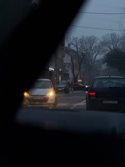A street scene is visible from inside a vehicle, partially obscured by the vehicle's frame. Traffic is lined up on a wet road. A white van with its headlights on is in the foreground, followed by a dark sedan. Pedestrians are crossing the street. Buildings are visible on the side of the road, along with bare trees against a grey sky, suggesting a cold or overcast day. Vehicle license plates are visible on the van and sedan.