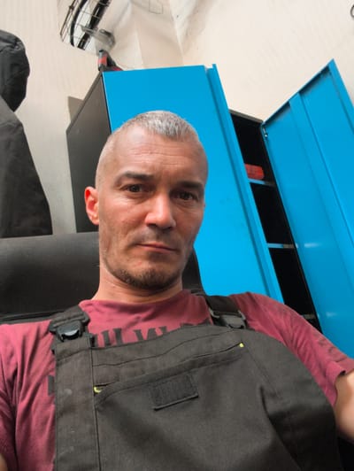 This is a selfie of a man with short grey hair and a light beard, wearing a maroon t-shirt and dark grey overalls. He is sitting in a black chair, with his upper body and face filling most of the frame. Behind him, there are bright blue metal cabinets with one door open, revealing black shelves and a red object. To the left, a dark coat hangs on a wall. The background suggests an indoor setting, possibly a workshop or garage, with fluorescent lighting visible overhead. The man's expression is neutral. The time of day and weather are not discernible. There is no visible text in the image.