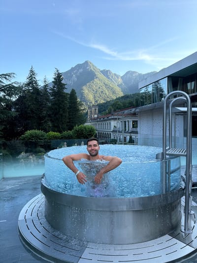 In San Pellegrino Terme, Italy, a man relaxes in a clear-walled, circular hot tub filled with bubbling water. The hot tub is situated on a tiled patio with a metal grate around its base. A metal ladder with two steps and a handrail is attached to the side of the tub. In the background, a modern building with large glass windows stands adjacent to older, traditional Italian architecture. Beyond the buildings, lush green mountains rise under a clear blue sky with a crescent moon visible. Trees line the landscape between the hot tub and the buildings. The man, with dark hair and a beard, is smiling at the camera, his arms resting on the edge of the tub. Water splashes around him.