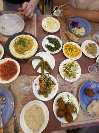 A dining table in a restaurant, located in the Galil Elyon Regional Council, Israel, is depicted from a top-down perspective. The table is covered with patterned brown paper placemats, one of which displays the Hebrew text "גליל עליון" (Upper Galilee). The table is densely covered with approximately sixteen small dishes containing a variety of Middle Eastern food.
Key food items include:
* A large black plate of hummus topped with stewed chickpeas and fava beans (ful medames) and garnished with parsley.
* Small white plates holding shredded white cabbage salad, finely chopped green herbs (possibly tabbouleh components), corn salad with diced peppers, mashed eggplant salad, pickled cucumbers, and tomato salsa.
* A small blue plate with mixed pickled cabbage.
* Another small blue plate with one falafel ball, a piece of pita bread, and a white smear of sauce.
* A white plate with four falafel balls, garnished with lettuce and shredded carrot.
* A white plate containing a thick, light-colored dip, likely tahini or baba ghanoush.
* A white plate with a cooked vegetable and meat mixture.
* An additional white plate with shredded cabbage salad.
* One empty white ceramic plate is visible on the upper left, containing a few pistachio shells.
Two individuals are partially visible: a person's forearm and hand with red nail polish and a red bracelet are on the upper left of the image, resting near an empty plate. Another person's forearm and hand, wearing a white shirt, are visible on the upper right. Two empty clear drinking glasses are present, one inverted. Metal forks and a knife are positioned around the plates.