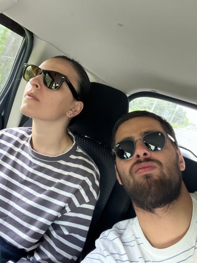 Two individuals, a woman and a man, are seated in the interior of a vehicle. The woman, positioned on the left, is wearing a striped t-shirt and sunglasses, with her head tilted upwards and her gaze directed out the window. She has dark hair pulled back and multiple earrings in her left ear. The man, on the right, is also wearing sunglasses and a t-shirt with subtle stripes. He has dark hair and a beard and is looking towards the camera. The interior of the car appears to be a standard passenger vehicle, with dark seats and a light-colored headliner. Through the windows, a blurred view of greenery and what appears to be a building or structure is visible, suggesting an outdoor environment. The image is a selfie-style shot taken from within the vehicle. The location is not discernible from the image itself, and no information about Achkhoti, Georgia can be inferred.