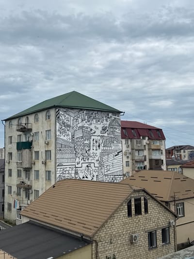 The image displays a cityscape with a focus on apartment buildings under a cloudy sky. The buildings are varied in architecture and age, with some featuring older, stucco facades and others having more modern brickwork or corrugated metal roofing. A striking feature is a large black and white mural covering a significant portion of one building's facade. The mural depicts a chaotic, busy scene with numerous small figures engaged in various activities, as well as representations of buildings and everyday objects.
The perspective is from a slightly elevated viewpoint, looking up at the buildings and the sky. The sky is overcast with soft, diffused light, suggesting it is either early morning or late afternoon, or simply a cloudy day. The overall mood is somewhat somber due to the weather, but the vibrant mural adds a touch of life and visual interest to the urban environment.
The image appears to be taken in Derbent, Russia, as indicated by the prompt. There is no discernible activity or event taking place in the immediate scene, but the mural itself suggests a narrative or commentary on urban life. Visible text is limited to a small signature or artist's mark on the mural itself, which is too small to be legible in the provided image. Other details include numerous air conditioning units mounted on the building walls and various windows, some with balconies.