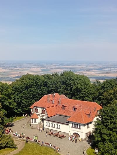 This is a high-angle, full-shot, medium-distance, exterior view of a building with red-tiled roofs, featuring white walls and dark timber-framed accents, situated in a rural landscape. The building is a guesthouse or restaurant located on a hilltop, indicated by the expansive view of fields and distant haze. The foreground shows a paved courtyard with scattered patrons sitting at picnic tables and on benches. There are several people walking and gathered in small groups. The building has multiple windows and dormer windows protruding from the roof. Surrounding the building and courtyard are lush green trees, and a grassy slope is visible in the lower left corner. The sky is a clear, pale blue, indicative of a sunny day. The building appears to be on Mount Ślęża in Sobótka, Poland.