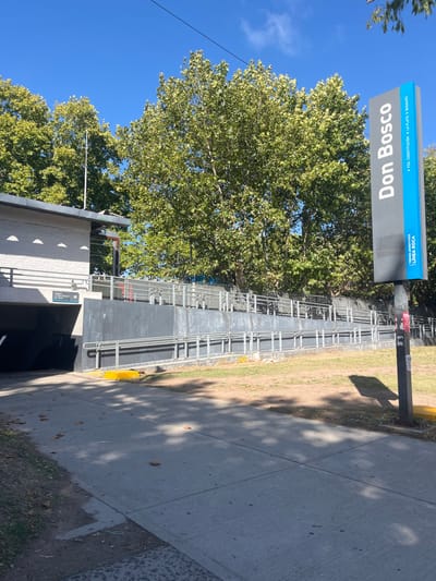 The image shows the Don Bosco train station entrance on a sunny day. A blue sign with white lettering displays "Don Bosco" above an entrance structure with gray walls and silver railings. A large tree with green leaves obscures part of the station. A concrete sidewalk runs along the right side, and grass and greenery fill the adjacent area. Shadows from the tree are cast on the sidewalk.