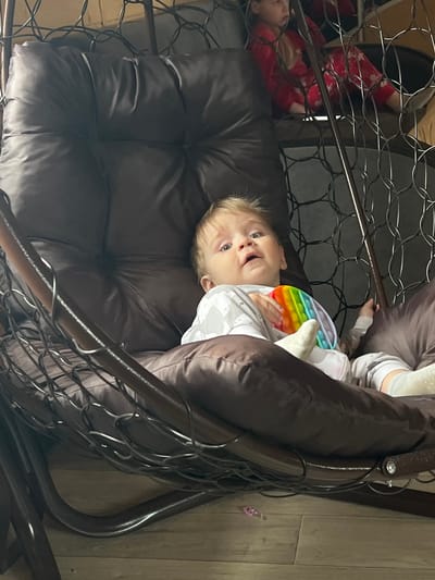 A toddler is lying in a brown cushioned chair that is enclosed by a dark metal frame, possibly a hanging egg chair. The child, wearing a light-colored patterned pajama set and white socks, is holding a rainbow-colored sensory toy in their hands. Another child, dressed in red pajamas with white snowflake patterns, is visible in the background partially obscured by the chair's metal frame. The setting appears to be indoors, with a light-colored wooden floor visible in the background. The location is Bol'shakino, Russia, but there are no landmarks to confirm.