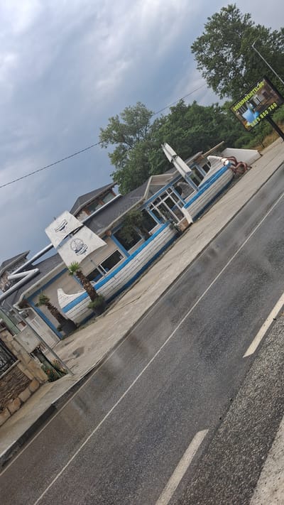 The image shows a building designed to resemble a ship, located in the city of Marchevo, Bulgaria. The building is white with blue trim and has a ship's hull as its main structure. It features a sail-like design element on the roof. Palm trees are planted in front of the building. A road with white lines is visible in the foreground, and the ground appears wet. A billboard for "GREENPARKHOTEL.BG" with a phone number is visible in the background. The sky is overcast.