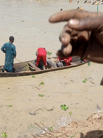 The scene, captured in Kokani North, Nigeria, features individuals engaged with a murky brown body of water. In the midground, a weathered wooden dugout canoe is partially beached in shallow, muddy water adjacent to a dirt bank. Three adult males are visible in and around the boat: one standing at the stern, facing away, clad in a dark teal long tunic and holding a pole; a second, wearing a red shirt and red pants, is bent over inside the boat; and a third, in reddish attire, is seated or crouching in the boat, near a visible fishing net. The immediate water surface is dotted with small green aquatic plants and floating debris. The background reveals a broader expanse of the brown water, populated by multiple distant figures partially submerged, their heads and upper torsos visible above the surface, indicative of wading or swimming. A yellow float is discernible among these figures. The obscured foreground on the right presents a dark-skinned human hand, out of focus, with fingers slightly curled.