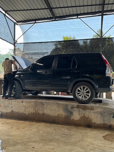A black SUV is parked under a metal awning in Matangkeupala Sa, Indonesia. The vehicle is parked close to a concrete wall. A man wearing a beige shirt, black pants, and sandals is standing near the front of the SUV with the driver's side door open. The environment includes metal awning structures, trees, and a concrete wall.