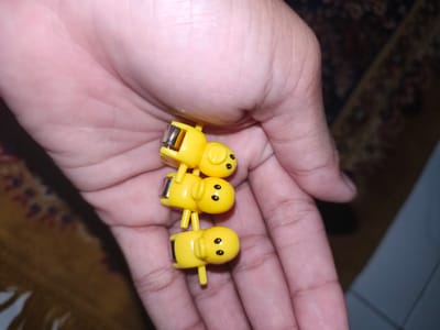 The image displays a human hand holding three yellow, pig-shaped figurines. The figurines are stacked vertically, with each pig appearing to be a miniature toy. They have simple, cartoonish features with black eyes and a small snout. A metallic component is visible on the back of each figurine, suggesting they might be decorative clips or accessories. The hand is positioned in the center of the frame, with the palm facing upwards and fingers slightly curled. The background is out of focus, showing a patterned carpet and a dark surface, likely a floor. The lighting appears to be indoor. The image was captured in Barengkrajan, Indonesia.
