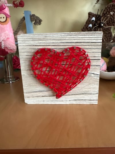 A close-up, eye-level shot shows a rustic wooden board with a red string art heart as the central subject. The heart is constructed from numerous strands of red yarn stretched between small metal nails hammered into the board. The wooden board has a weathered, whitewashed plank appearance. To the left of the board, partially visible, is a pink doll or craft with lace trim and a large button with two holes. Behind the board and to the right, a brown crocheted figure with googly eyes and antennae is visible, resembling an insect or animal. In the background, a light yellow wall is present, and a white bowl containing colorful decorative items, including what appears to be a pink glittered sphere, is situated to the right of the wooden board. The objects are placed on a brown wooden surface, likely a table or shelf. The image composition focuses on the handcrafted heart. The location is not determinable from the image.