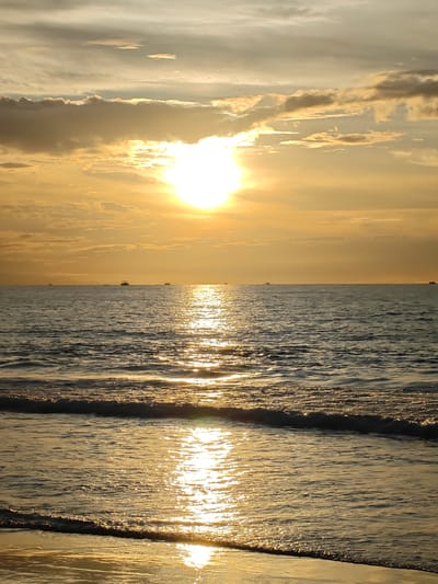 The image depicts a serene seascape at sunset, likely in the vicinity of Peuneulet Baroh, Indonesia, given the context. The sun, a bright, luminous orb, hangs low in the sky, casting a warm, golden glow across the scene. Its light reflects brilliantly on the water's surface, creating a shimmering pathway that extends towards the horizon.

The sky is adorned with a mixture of clouds. Some are wispy and thin, allowing the sun's rays to penetrate, while others are more dense and textured, adding depth and drama to the celestial canvas. The overall color palette of the sky is dominated by warm oranges, yellows, and muted grays.

The sea itself is calm, with gentle waves rolling towards the shore. The water appears dark and deep, contrasting with the bright reflections of the sun. In the distance, several small boats are visible, silhouetted against the horizon, suggesting a tranquil maritime activity.

The foreground of the image shows a sandy beach, wet from the receding tide. The wet sand also catches the golden light, appearing smooth and reflective. The edge of the water gently laps at the shore, forming a fine line of foam. The overall impression is one of peace and natural beauty, characteristic of a coastal sunset. There are no people or specific identifiable structures that indicate a bustling urban environment.