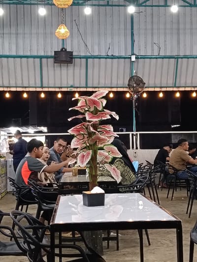 The scene depicts an active, semi-outdoor dining establishment at night, located in Namplohbaro, Indonesia. The overhead structure features a corrugated metal roof supported by green metal beams. Illumination is provided by strings of exposed white light bulbs and two decorative hanging wicker pendant lamps. A black speaker is mounted on a beam to the left, and a black wall-mounted fan is visible on the right.
In the foreground, a rectangular table with a white marble-patterned top and a black metal frame is present. A black tissue box rests on this table. Dominating the midground is a tall artificial plant with numerous large pink and green variegated leaves, resembling a Caladium.
To the left of the plant, three individuals are seated at a table. A young male in a grey and orange short-sleeved shirt is holding and looking at a smartphone. Adjacent to him, a female wearing a light brown hijab is partially visible, also looking at the phone. An adult male with eyeglasses, wearing a dark grey shirt, is seated beside them, leaning forward and observing the phone screen. Drinks in glasses are visible on their table.
To the right of the plant, additional patrons are seated at dark tables with black chairs. One male, wearing a brown t-shirt and jeans, is seated and appears to be looking at a phone. Another male in a black t-shirt is partially visible behind him. A female in a black and yellow patterned garment is partially visible behind the plant.
In the background on the left, a person in a dark blue shirt stands behind a counter where white objects are displayed. The floor throughout the establishment appears to be a light-colored, possibly concrete or packed earth, surface.