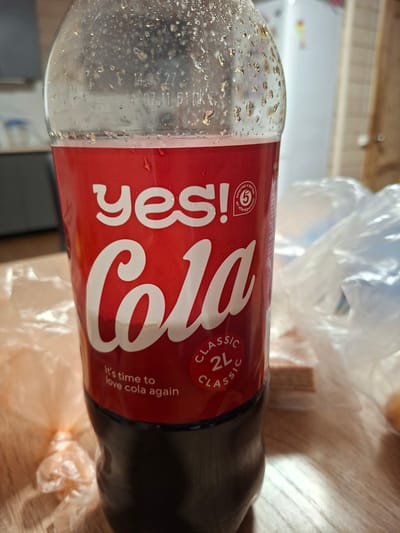A close-up, eye-level, medium shot captures a 2-liter bottle of "Yes! Cola" with a red label, condensation on the bottle, and the drink itself being a dark brown liquid. The label features the brand name "yes!" in white, followed by "Cola" in large white script. Circular emblems are present on the label, with one showing the number "5" and text in Cyrillic. Another circular emblem contains the text "CLASSIC 2L CLASSIC". Below the "Cola" script, the phrase "It's time to love cola again" is visible. The bottle is placed on a wooden surface, and behind it, plastic bags and a blurred background of what appears to be a domestic interior are visible. The bottle likely contains a cola-flavored carbonated beverage. The image was taken indoors, possibly in a kitchen or dining area. There are no people visible in the frame. The lighting appears to be artificial, casting soft shadows. The scene is static, with no discernible action. This image does not provide any information to determine its location in Medvezhyi Ozyora, Russia.