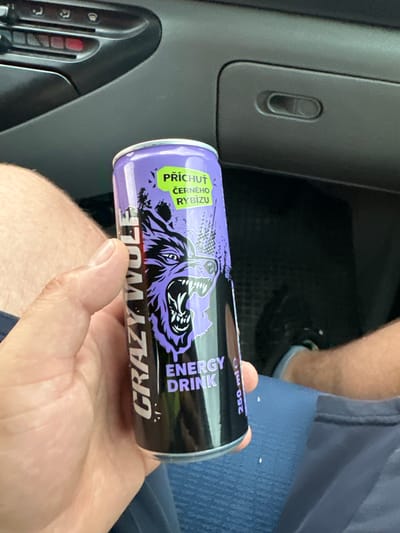 A person holds a can of "Crazy Wolf" energy drink, displaying a purple and black design with a wolf's head graphic. The can is oriented horizontally, with text visible on its surface. Text on the can includes "PŘÍCHUŤ ČERNÉHO RYBÍZU" (Flavor Blackcurrant) in a green box and "ENERGY DRINK" vertically oriented. The brand name "CRAZY WOLF" is also prominently displayed, though parts of it are obscured by the hand holding the can. The background shows a car interior, with a dark gray dashboard and a blue fabric seat. A person's legs are visible in the background, wearing dark shoes. The scene appears to be taking place indoors, likely in a vehicle, given the dashboard and seat. There are no strong indicators of time of day or weather conditions.