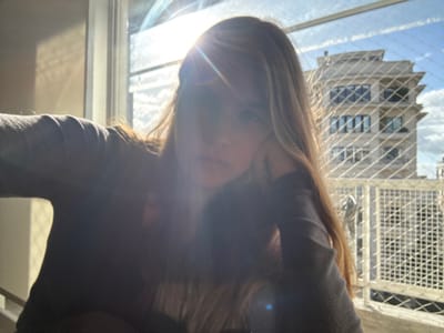 The image is a backlit, selfie-style close-up of a female subject positioned indoors, looking towards the camera through a window or balcony opening in Porto Alegre, Brazil. Intense sunlight from the upper left quadrant creates a prominent starburst lens flare, heavily obscuring the subject's face and upper torso, rendering her largely in silhouette. She has long, light-colored hair and is wearing a dark, long-sleeved garment. Her right hand appears to be resting near her face, possibly supporting her head.

The view beyond the subject is framed by a white, diamond-patterned safety net that covers the entire window or balcony opening. In the urban background, beneath a partly cloudy sky, a multi-story, light-colored building with a distinctive stepped or tiered architectural design is visible. The building features multiple windows and balconies, indicative of a residential or commercial structure typical of the city environment.