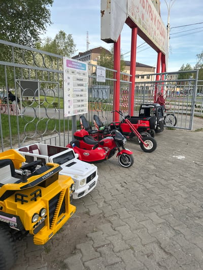 This image, taken at a sideways angle, shows a collection of children's electric ride-on vehicles lined up next to a gate. The vehicles, including yellow and white trucks, a red scooter, and a red ATV, are parked on a gray brick-paved area. A sign attached to the gate displays the operating hours of the "Culture and Recreation Park" from May 18th to May 24th. The sign indicates that the park is closed on Mondays and Tuesdays. The image includes a partial view of the gate and a building with multiple windows in the background. There is also a tree on the left side of the gate. The image location is identifiable as Uray, Russia, based on the Cyrillic text "ПАРКА КУЛЬТУРЫ И ОТДЫХА" (Park of Culture and Recreation) on the sign.