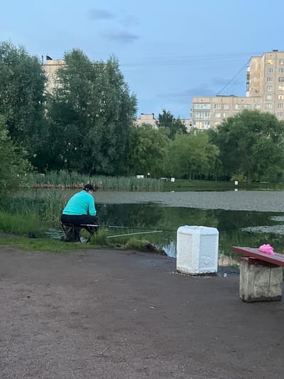 The image depicts a person fishing by a body of water, likely a pond or small lake, in Rahya, Russia. The setting appears to be an urban or suburban park area, with tall, apartment buildings visible in the background to the left. Green trees and reeds line the edge of the water. The time of day seems to be late afternoon or early evening, as indicated by the soft, diffused light. The sky is clear with some faint clouds.

The main subject is a person, seen from the back, wearing a bright teal shirt and dark pants. They are sitting on a chair or stool, facing the water, and appear to be actively engaged in fishing, with fishing rods visible. Beside the person is a fishing bag. A large white styrofoam cooler is placed on the ground near the water's edge, and a wooden bench with a pink object on it is visible to the right. The ground in the foreground is a dark, somewhat wet asphalt or gravel path. The overall atmosphere is calm and peaceful, suggesting a recreational activity. There is no visible text in the image.