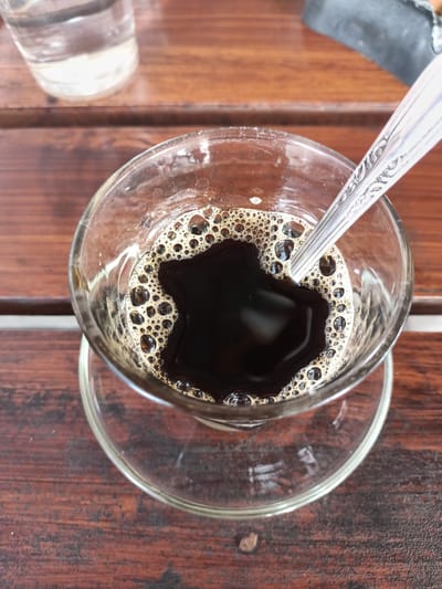The image captures a close-up, overhead view of a glass of dark coffee with a frothy surface, placed on a saucer. A silver spoon with an ornate design rests inside the glass, partially submerged in the coffee. The coffee is served on a wooden surface, likely a table, which shows a weathered texture and grain. In the background, out of focus, is a glass of clear liquid, possibly water. The overall setting appears to be a casual cafe or dining area. The image does not provide any specific details about its location in Cotgadong, Indonesia, or any people present.