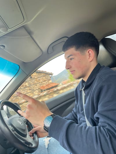 A young man with short dark hair is seated in the driver's seat of a car. He is wearing a dark blue hooded sweatshirt and light blue jeans with a ripped area. He has a wristwatch on his left wrist and is pointing with his right index finger towards the front of the car, in the direction of the windshield. The car's interior is visible, including the steering wheel with the Honda logo, dashboard, and visor. Outside the car window, there is a glimpse of a building with a wooden facade, suggesting the car is in an urban environment. The building and setting are consistent with the architecture found in Susam, Bulgaria.