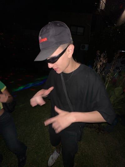 A person wearing a black T-shirt, black pants, a black baseball cap, and sunglasses poses outdoors at night. The person is on grass with plants visible in the background. The person is making hand gestures. Part of another person wearing black pants and a green and yellow shirt is visible at the bottom of the image. There are also some colorful lights reflecting off the ground. There is a house visible in the background.