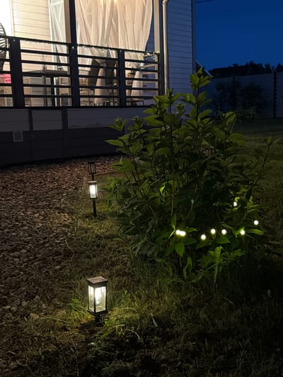 The image depicts a nighttime scene in the city of Alyoshkino DP, Russia. In the foreground, two solar-powered garden lights illuminate a patch of grass and gravel. One light is positioned closer to the viewer, while the other is further back, near a bushy green plant. This plant is adorned with small, glowing fairy lights that create a whimsical effect. In the background, a balcony or porch area is visible, enclosed by a dark railing and covered with white curtains. Furniture, including chairs and a table, can be seen through the railing. The overall atmosphere is serene and dimly lit, with the dark blue night sky visible above. There are no people present in the image.