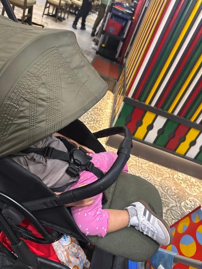 The image shows a child sitting in a stroller. The child is wearing a pink outfit with white hearts and white Adidas sneakers. The stroller is olive green and has a black harness. In the background, there are colorful vertical stripes and a patterned floor. The image appears to have been taken indoors, possibly in a public place like a mall or shopping center, given the stroller and the background elements that suggest a commercial environment. There are no visual cues in the image that can confirm or deny its location in Gunung Putri, Indonesia.