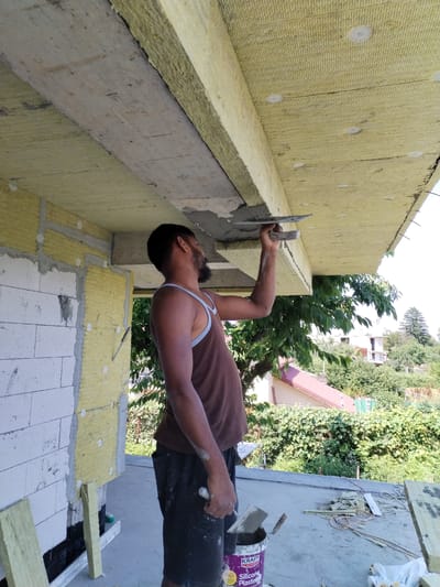 A construction worker is applying plaster to the underside of an exterior overhang. The worker, a dark-skinned man with short dark hair and a beard, wears a brown tank top and dark shorts. He holds a trowel in his right hand, applying plaster to a horizontal surface covered with yellow insulation panels. The background shows a residential area with houses and trees. The scene appears to be outdoors on a sunny day. The location is Dumbrăveni, Romania.