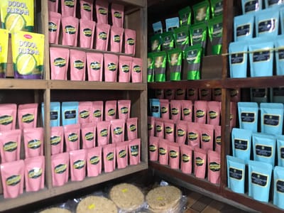 This indoor shot depicts a retail or storage space, likely a shop or stall in Panteepanah, Indonesia, showcasing various packaged food products neatly arranged on wooden shelves. The scene is dominated by a variety of goods, primarily in pouches and boxes.

On the left side of the image, the top shelf features yellow packages labeled "DODOL DURIAN" and "Sabila," with one clearly showing "BEST QUALITY" and an illustration of a durian fruit. Below these, and extending to the middle and lower shelves, are numerous small pink pouches. Many of these pink pouches have a black and gold label indicating "BEST QUALITY," and some specifically mention "ARABICA GRADE ONE KOPI GAYO" or "ARABICA GRADE ONE."

The right side of the image displays more shelves, holding a collection of green and light blue pouches. The green pouches also feature "BEST QUALITY" labels, while the light blue pouches often state "HIGH QUALITY" and identify the product as "ARABICA NATURAL GAYO" or "ARABICA NATURAL GAYO KOPI GAYO." The repeated text "BEST QUALITY" and "HIGH QUALITY" suggests a focus on premium products.

At the very bottom of the image, several round, flat, light-colored objects are visible, possibly crackers or other snacks, packaged in clear plastic. The arrangement implies an organized display for sale, presenting a range of local Indonesian products, prominently featuring durian confectionery and Gayo coffee.