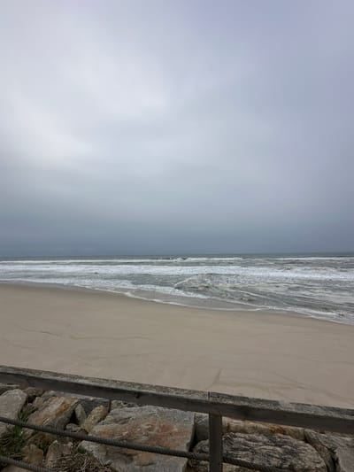 The image shows a beach in Vagos, Portugal. The sky is overcast and grey. The ocean is on the right side of the image with waves washing onto the sandy beach. A stone wall is on the right edge of the frame, with a wooden post and rope alongside it.