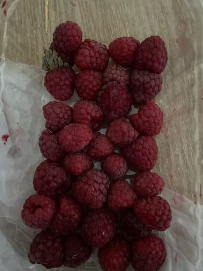A pile of ripe raspberries is contained within a clear plastic bag, which rests on a light-colored, textured surface. The raspberries are a deep red color and appear plump and juicy. Some of the berries have a light fuzz visible. In the upper left corner, a small cluster of dried leaves and stems is also present within the bag, mixed with the raspberries. The background of the image is slightly blurred but suggests a wooden or woven texture. There is no visible indication of the city of Bilohorodka, Ukraine, in this image.