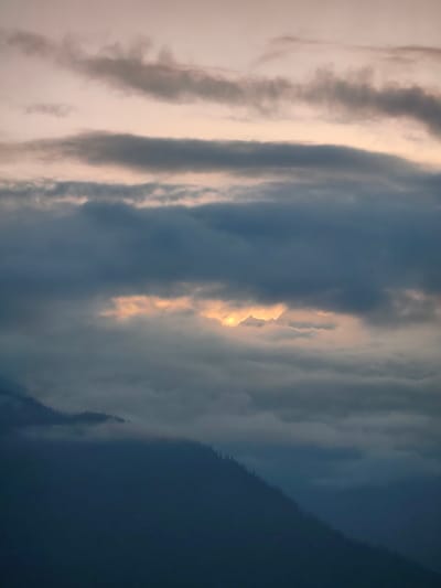 This is a vertical, low-angle shot of a mountainous landscape at dusk, likely in the region of Aleo, India. The sky is a pale pinkish-beige, with horizontal bands of dark gray clouds obscuring much of the view. A sliver of golden light breaks through the clouds in the midground, illuminating the peaks of jagged mountains. The lower portion of the image is dominated by a dark, mist-shrouded mountainside, with a sparse scattering of pine trees visible on the upper ridge, their silhouettes appearing as dark spikes against the muted sky. The overall atmosphere is somber and serene.