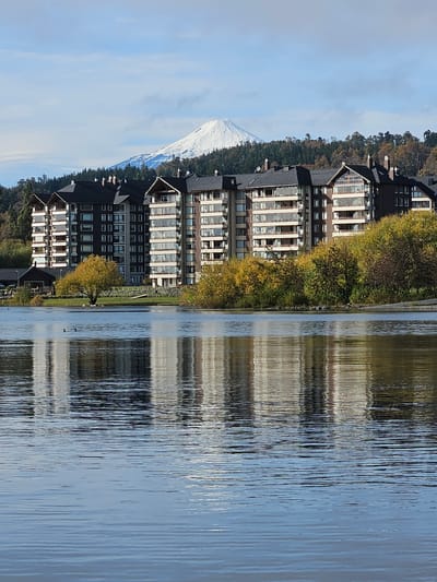 The image depicts a lakeside scene in Villarrica, Chile. A multi-story building with numerous windows stands adjacent to the lake's edge. The building's reflection is visible in the water, which has a textured surface due to wind or ripples. Trees with autumn foliage are present along the shoreline, adding color to the scene. In the distance, a snow-capped mountain is visible against a blue sky with clouds. Two ducks are seen in the lake near the shore.