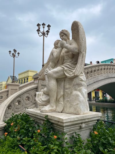 The image depicts a marble sculpture of two figures, likely an angel and a child, situated in front of a body of water. The angel, with large wings, is seated and appears to be embracing the child. The sculpture rests on a marble pedestal adorned with classical motifs. Behind the sculpture, a bridge with intricate stonework arches over the water. On the bridge and in the background, several people are visible, as well as ornate lampposts and buildings with yellow and green facades. Lush green bushes with orange flowers surround the base of the sculpture and the water's edge. The sky is overcast. This scene is located at the Hoi An Impression Theme Park in Nghĩa Trụ Commune, Vietnam, a place designed to evoke a sense of historical European charm.