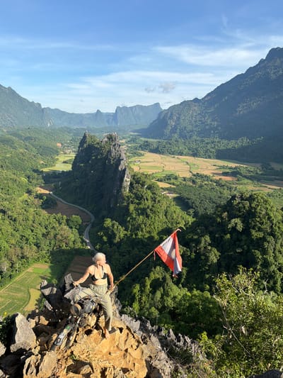 The image depicts a scenic mountainous landscape in Naka, Laos, under a bright, clear sky with wispy clouds. In the foreground, a person with blonde hair is seated on a rocky outcrop, holding a flag on a pole. The person is wearing a black bikini top and olive green pants. Next to them is a vintage black motorcycle. The flag is red, white, and gray. The rocky outcrop is surrounded by lush green vegetation and overlooks a valley with rice paddies and a winding road. In the background, more rugged mountains rise, covered in dense green forest. The overall scene captures a sense of adventure and exploration in a natural, mountainous setting.
