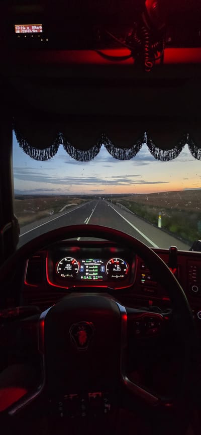 The image, captured from the perspective of a truck driver, shows a highway stretching into the distance during either dawn or dusk. The dashboard of the truck is illuminated with red lighting, displaying various metrics including speed (89 km/h) and time (07:14). The road ahead is paved with white lines, and the surrounding landscape is dry and sparsely vegetated. The sky transitions from a pale blue at the top to hues of orange and pink closer to the horizon, indicating a sunrise or sunset. The truck's interior is dark, with the exception of the illuminated dashboard and red ambient lighting. A decorative fringe hangs from the top of the windshield. The location is stated to be the city of Calatorao, Spain.