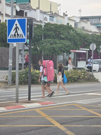 A pedestrian crossing on a sunny day in Sant Andreu de Llavaneres, Spain, shows two adults and a child walking across a road marked with yellow grid lines and a red and white curb. The older adult in the foreground is carrying a large, pink bag. A pedestrian crossing sign is visible, with a green traffic light illuminated. Buildings and vegetation line the background. A bus stop with seated individuals is visible in the midground.