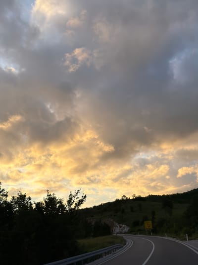 This media file presents a scenic landscape in Jasenovo Polje, Montenegro, captured at either sunrise or sunset, indicated by the dramatic lighting. The image is dominated by a expansive sky featuring a striking contrast: the upper left portion is filled with dark, grey storm clouds, while the lower right is illuminated by warm, golden-orange light, highlighting the edges and undersides of dense cloud formations.

Below the dynamic sky, a verdant, tree-covered hillside stretches across the lower right, appearing mostly in shadow. The trees along the ridge are silhouetted against the brighter sky, while those closer to the foreground show darker green tones. A distinct rocky outcrop with lighter stone is visible on the hillside.

A paved road with white lane markings and a metal guardrail curves along the far right side of the frame, winding through the hilly terrain. On the hillside adjacent to the road, a yellow diamond-shaped road sign is visible. The text "JASENOVO POLJE" is clearly discernible on the sign, accompanied by an arrow pointing upwards, indicating a directional cue for the road ahead. Another arrow points left, likely to a different destination, but the associated text is not fully legible. No people or specific activities are depicted, emphasizing the natural beauty and serene atmosphere of the mountainous region.