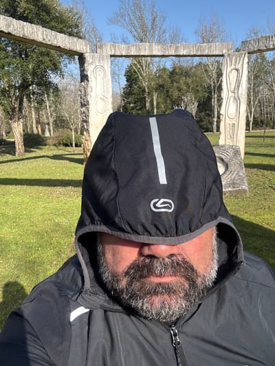 The image presents a close-up, chest-level view of an individual standing outdoors. The subject's face is partially obscured by a black hooded jacket with a reflective vertical stripe and a "Kalenji" brand logo on the hood. A thick, salt-and-pepper beard covers the visible lower face, including the chin and cheeks. The jacket is zipped.
In the background, under a clear blue sky, an outdoor art installation is visible. It comprises several large, rough-hewn concrete or stone structures forming an archway. These structures feature abstract, carved designs; a prominent vertical slab displays a spiral-like motif, and another partially visible structure to the right contains a circular indentation. The immediate environment consists of green grass and numerous trees, some with bare branches, suggesting a natural park or forest setting under bright daylight.
This scene is consistent with the "Monumento à Batalha do Buçaco" (Monument to the Battle of Bussaco) located within the Mata Nacional do Buçaco (Bussaco National Forest) in Luso, Mealhada, Portugal.