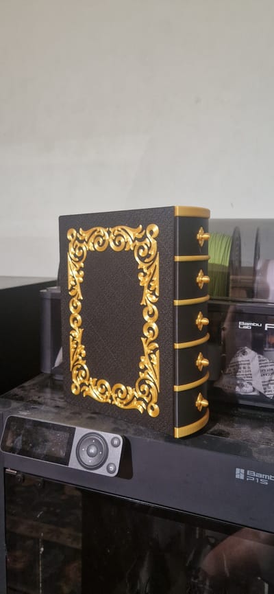The image shows a close-up of a 3D-printed object resembling an ornate book, placed on top of a black electronic device. The book has a textured dark gray cover with an intricate golden filigree design framing a central rectangular area. The spine of the book is also dark gray with golden decorative elements that mimic raised bands and studs.

The electronic device appears to be a 3D printer, indicated by the visible branding "Bambu P1S" on its surface. The printer has a control panel with a circular navigation button and a small screen. In the background, out of focus, are spools of filament, likely for the 3D printer, one of which is bright green. There are also some white bags or packaging in the background, one with visible text including "Bambu Lab". The overall environment suggests a workshop or crafting space. The image is taken from a slightly elevated angle, looking down and to the right.

The location of South Holland, United Kingdom, is not depicted in the image.