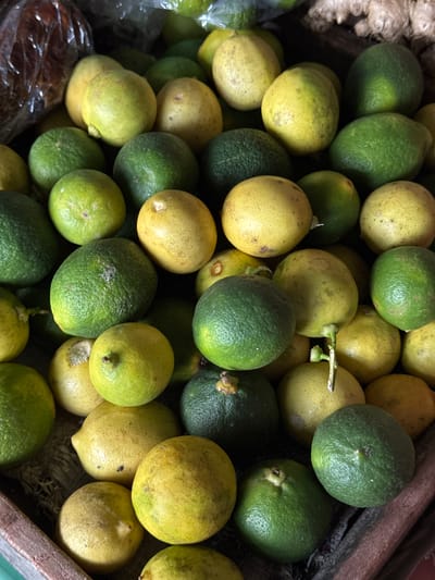 A top-down, close-up shot shows a large pile of lemons and limes in a wooden crate. The fruits are a mix of bright yellow and dark green, with some showing slight imperfections such as browning or small blemishes. The crate appears to be made of rough wood and is filled with what looks like straw or dried grass at the bottom. In the upper left corner, a portion of a plastic bag with a crinkled texture is visible. The lighting is subdued, creating shadows that highlight the rounded shapes of the citrus. The scene is devoid of any human presence. The location is identified as Karangsambung, Indonesia.