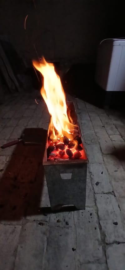 An outdoor scene at night shows a rectangular metal brazier filled with glowing coals and flames. The brazier is situated on a paved surface, possibly a patio or courtyard. To the left of the brazier, a metal spatula with a red handle rests on the ground, casting a shadow. In the background, a dark, nondescript structure is visible, and to the right, a portion of a white appliance, possibly a washing machine, can be seen. The overall impression is of a domestic outdoor space at night, with a fire being prepared or maintained. The location in Andijani, Uzbekistan is not explicitly depicted in the image but is provided as context.