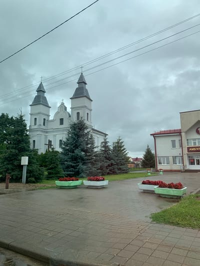 The image captures a wide-angle view of a church and a building with Cyrillic lettering reading "АМБУЛАТОРИЯ" (Ambulatory) in the village of Сноўскі сельскі Савет, Belarus. The sky is overcast and gray, with power lines stretching across the upper portion of the frame.

In the foreground, a paved area with a tiled pattern is visible, wet from rain, with a curb and a strip of grass on the right. Several large, rectangular planters filled with red flowers are placed on the paved area.

To the left of the planters, a large, light-colored church with two prominent bell towers is the main architectural feature. The church has a cross on each tower and a larger cross in front of it. Evergreen trees, including tall fir trees, surround the church. A small, white rectangular object on a pole and an old-fashioned water pump are also visible near the church.

To the right of the church is a lighter-colored building with a red roof. This building appears to be an outpatient clinic or medical facility, as indicated by the "АМБУЛАТОРИЯ" sign. The building has multiple windows and an entrance with stairs.

The overall scene suggests a quiet, possibly damp day in a rural Belarusian settlement, with a prominent religious building and a public service facility.