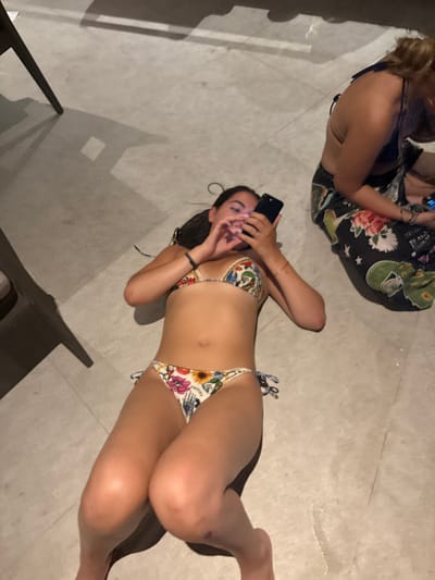 The image shows two young women in a tiled room with a grey floor. One woman is lying on her back and looking at a mobile phone. The other woman is sitting with her legs crossed to the side, looking down at her own phone. The woman lying down is wearing a colorful floral bikini, and the woman sitting down is wearing a black and floral sarong-like garment. The room is sparsely furnished, with only a piece of furniture out of frame in the bottom left corner. The coordinates provided place this scene in Santa Fe, Costa Rica.