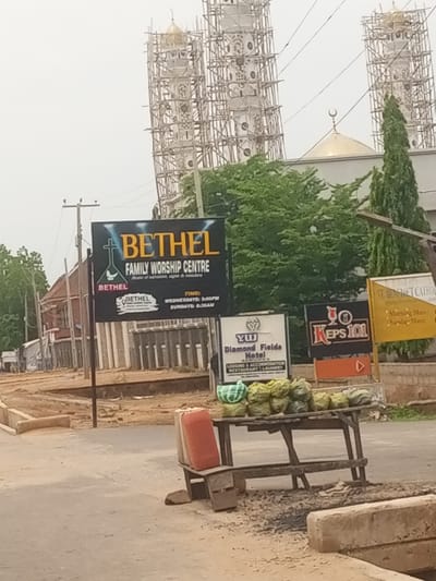 The image depicts a street scene in Baccure Kofare, Nigeria. In the background, two large towers, possibly minarets of a mosque, are under construction, encased in scaffolding. A golden dome is visible between them, topped with a crescent moon symbol. In the foreground, a prominent black sign advertises "BETHEL FAMILY WORSHIP CENTRE," detailing service times as Wednesdays at 5:00 PM and Sundays at 8:30 AM. Below this, another sign for "Diamond Fields Hotel" lists services including lodging, accommodation, restaurant, and laundry. A third sign, partially obscured, reads "KEPS 101." A yellow sign to the right mentions "ST. BENEDICT CATHOL" and "Morning Mass / Sunday Mass."

In the mid-ground, a wooden cart is laden with watermelons, some wrapped in green netting. A red container sits on a makeshift stool beside the cart. The environment appears to be dusty and somewhat underdeveloped, with dirt roads and construction evident. The sky is overcast, suggesting a cloudy or hazy day. The overall impression is of a commercial and religious area in a developing part of Nigeria, with ongoing construction and local commerce taking place.