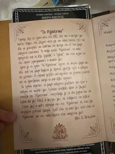 The image shows an open book or brochure laid flat, with a person's thumb visible at the bottom right corner, suggesting it is being held or pointed at. The page features Greek text with decorative borders and a stylized acanthus leaf motif.

The main text is titled "Το Ρεμούτσικο" (To Remoutsiko) and tells the story of its origins. It describes how, in 1982, Kostas Dimas, the father, transformed his father's storage room into a small tavern. The name "Remoutsiko" is explained as coming from a slang word used by the grandfather. Over time, the tavern grew, and its homemade dishes became popular, attracting customers from various areas. The text concludes with Ilias & Panagiotis, presumably the current operators, affirming their continuation of the legacy with the same passion and love taught by their father, aiming to offer a "delicious culinary experience."

To the left of the main text, a partial label or cover is visible with "ΚΤΗΜΑ ΛΑΝΤΙΔΗ - ΛΕΥΚΟΣ ΞΗΡΟΣ ΜΙΚΡΗ ΚΙΒΩΤΟΣ" (Lantidis Estate - Dry White Little Ark), indicating a connection to a Greek winery, likely in Agios Dimitrios, Greece. Another partial text on the right edge mentions "τα χαρακτηριστικά αρώματα της ποικιλίας όπως φύλλα ελιάς, τομάτα και ανθικές νότες" (characteristic aromas of the variety such as olive leaves, tomato and floral notes), further suggesting a wine or food product description. The scene appears to be indoors, under good lighting.