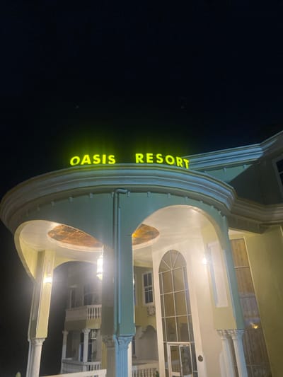 The image shows the facade of the Oasis Resort, located at the coordinates 4.145, 9.23 in the city of Buea, Cameroon. It is a nighttime shot of a building illuminated by artificial light. The building is mostly white with green accents, and features a large archway and a sign with the words “OASIS RESORT” in yellow letters. The sign is located above the archway, which is flanked by pillars. There is another archway visible inside the building. The building is lit by several lights, including a chandelier hanging from the ceiling of the inner archway and a pair of sconces flanking the outer archway. The image is taken from a low angle, looking up at the building. The sky is dark and there are no visible people or objects in the background. The building is surrounded by foliage. The overall feel of the image is one of tranquility and peace.