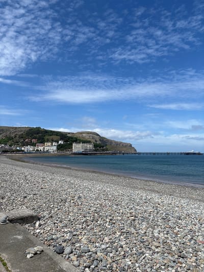The image shows a pebble beach in Llandudno, United Kingdom, under a blue sky with scattered clouds. The beach is composed of small, gray pebbles. In the distance, a mountain slopes down towards the coastline, with buildings situated along its base and on the lower slopes. A pier extends into the sea. The water appears calm. A concrete barrier runs along the edge of the beach in the foreground.