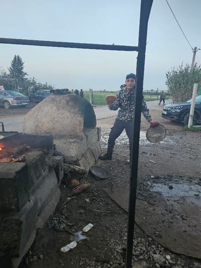 A young man stands near a large, dome-shaped oven, holding a round piece of bread in one hand and a cooking pan in the other. He wears a patterned jacket, dark pants, and a cap. The oven sits next to a muddy ground with scattered debris. A road with parked cars and green fields stretch in the background. A dark metal structure frames the image on the left side.