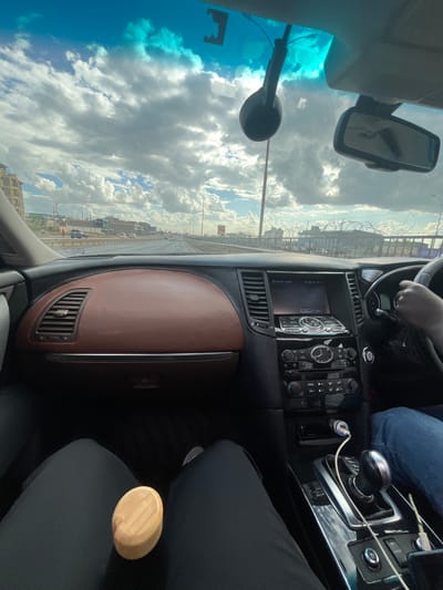 The image captures a perspective from inside a car, looking forward through the windshield and across the dashboard. The car appears to be traveling on a multi-lane road during the daytime under a bright sky filled with large, distinct cumulus clouds. The windshield has a bluish tint, and a rearview mirror is visible at the top, reflecting an indistinct shape. A dark, spherical object attached to a pole (likely part of the car's interior or an accessory) is prominent in the foreground near the windshield.

Outside the car, traffic can be seen in the distance on the road, which is flanked by a barrier topped with what appears to be barbed wire. Beyond the barrier, some buildings are visible, suggesting an urban or peri-urban environment.

Inside the vehicle, the dashboard features a two-tone design with a brown upper section and a darker lower section, including air vents. The center console houses an infotainment system with a screen that partially displays "Bluetooth Audio," surrounded by various buttons and knobs for controls. A white charging cable is plugged into a USB adapter near the gear shifter.

A driver's hand is visible on the steering wheel, and their legs, clad in blue jeans, are seen. In the front passenger seat area, a person's lap, covered by dark clothing (possibly black pants), holds a light-colored, oval-shaped wooden object, possibly a smooth stone or a small wooden box. The setting indicates an ordinary day of travel in Theta ward, Kenya, with clear weather conditions.