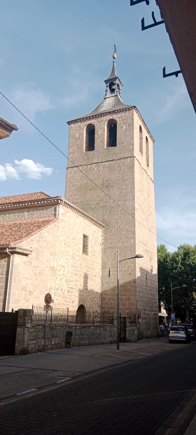 The image shows a stone bell tower of a church in Galapagar, Spain, with a clear sky overhead. The tower is tall and square, with arched openings where bells would be housed. A smaller, tiled roof structure is attached to the left of the tower. In front of the church, there is a stone wall with a black metal fence. A street runs in front of the church, with parked cars and a street light. A woman is walking on the sidewalk. The overall scene is quiet and appears to be in a well-maintained urban environment.